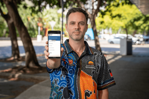 A man looking at the camera with a phone in his hand. The phone shows the service.nt.gov.au website. 